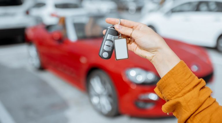 A white hand holding up a set of car keys in front of a red rental car.