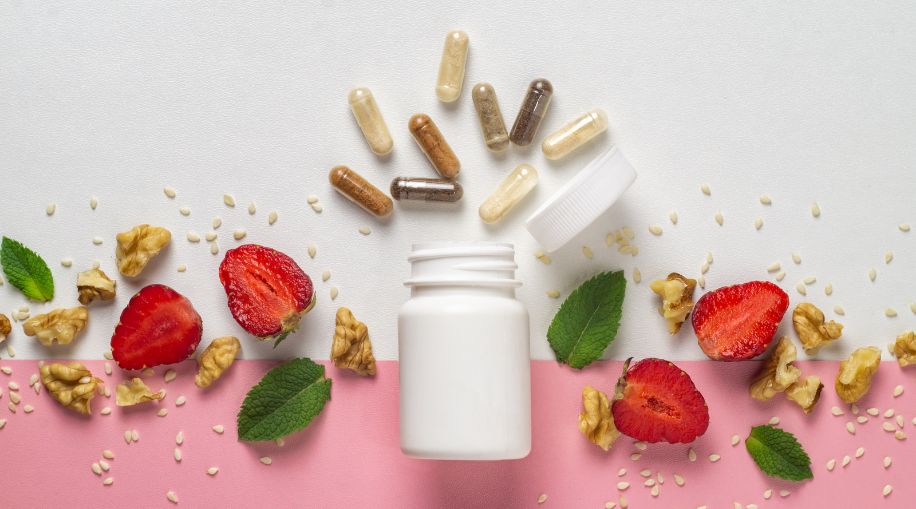 A pill bottle open on a table with vitamin capsules spilling out of it. There are strawberries and walnuts on the table as well. The background is white and pink.