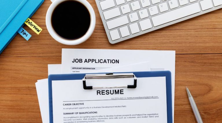 A resume attached to a clipboard on a table, below a mug of coffee and a keyboard.